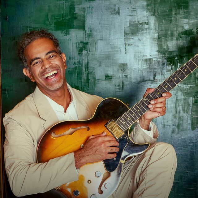 Jair Oliveira laughing while playing a hollow-body electric guitar
