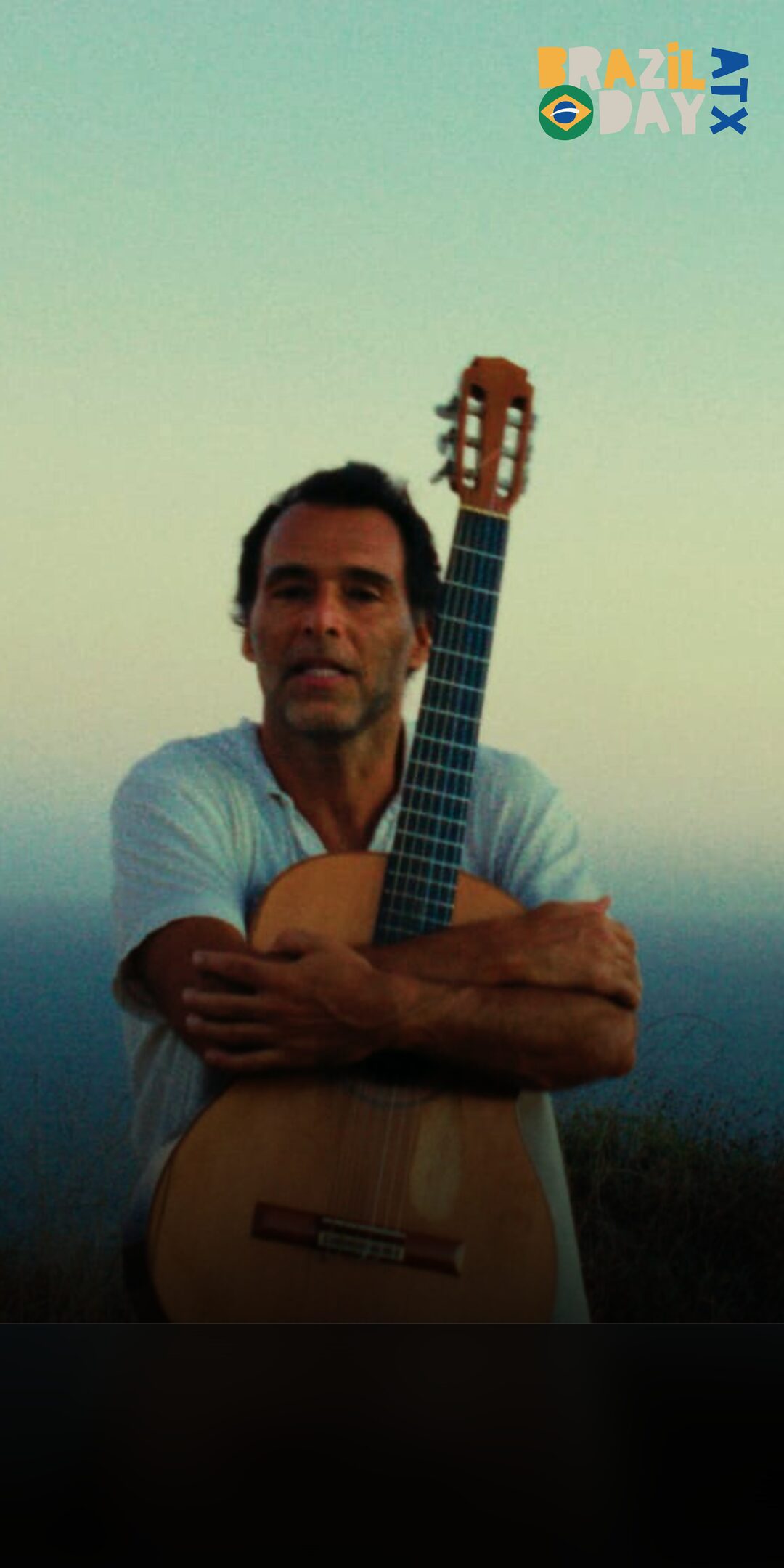 Rogê holding a nylon-string guitar at sunset by the sea