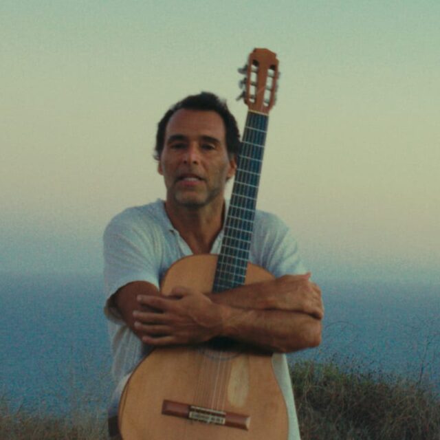 Rogê holding a nylon-string guitar at sunset by the sea